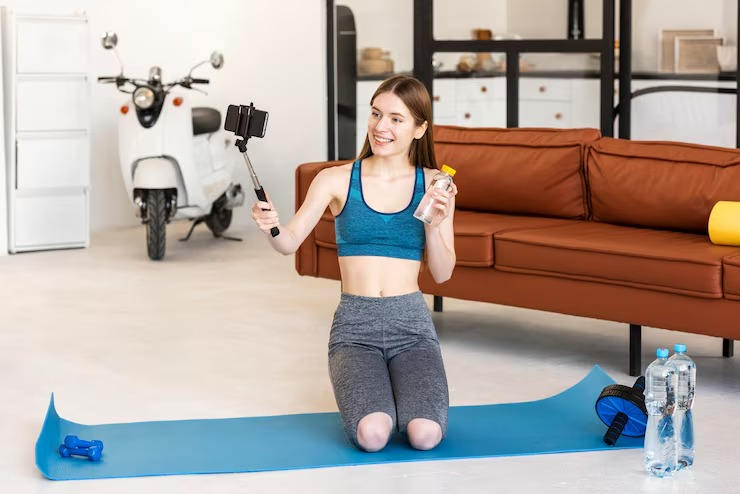 Fitness influencer kneeling on a yoga mat, filming herself with a phone and holding water.